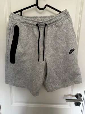 Grå Nike shorts med snörning - Snygga grå shorts från Nike med svart logga på vänster ben och svarta snören i midjan. De har elastisk midja och en svart detalj vid fickan. Perfekta för en avslappnad stil och tillverkade i mjuk bomullsmix.