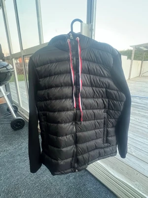 Svart moncler cardigan  - Snygg svart dunjacka från Moncler med quiltad design och huva. Jackan har dragkedja framtill, två fickor och coola röda och vita detaljer på snörena. Perfekt för kalla vinterdagar och riktigt stilren look.