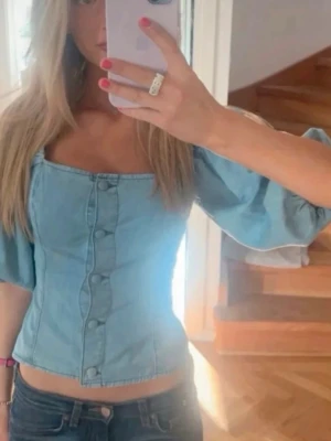 Ljusblå korsettopp med puffärm - Trendig ljusblå korsettopp i denim-look med puffiga trekvartsärmar och knappar framtill. Storlek S Snygg fyrkantig urringning och figurnära passform som framhäver midjan. Perfekt för dig som vill sticka ut med en cool och feminin stil.