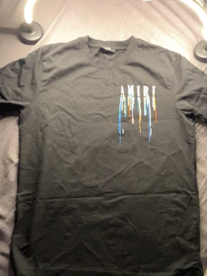Svart AMIRI t-shirt med färgstänk - Säljer en svart t-shirt från AMIRI med coolt tryck framtill där loggan har färgstänk i blått, vitt, orange och gult som rinner ner. Klassisk passform och rund hals. Perfekt för dig som vill ha en unik och trendig look.