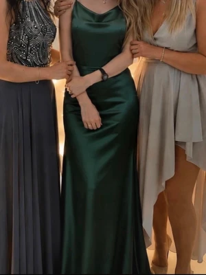 Grön långklänning i satin - Mörkgrön långklänning i glansigt satinmaterial med smala axelband och elegant draperad halsringning. Klänningen har en slank och figurnära passform som ger en lyxig känsla. Perfekt för dig som vill sticka ut med en stilren och trendig look.Haft på mig 1 gång ,inga fläckar eller skador. Den kan ha i vissa ställe glitter vilket går bort med hand.