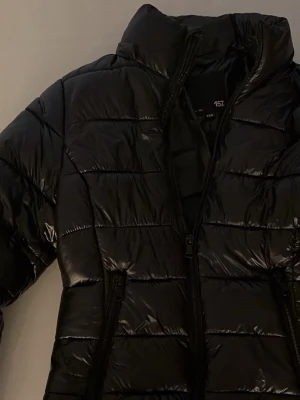 glansig pufferjacka - Säljer en svart, glansig pufferjacka med dragkedja och hög krage. Jackan har en klassisk quiltad design och är perfekt för kalla vinterdagar. Passar dig som vill ha en stilren och trendig look. Materialet är syntet och jackan är vadderad för extra värme. pris går att diskutera
