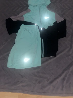 Nike Running set i mintgrönt - Nike Running set med jacka och shorts i mintgrönt med svarta och grå detaljer. Jackan har huva, dragkedja och reflekterande logga. Shortsen har resår i midjan och reflekterande detalj. Perfekt för löpning eller träning utomhus.