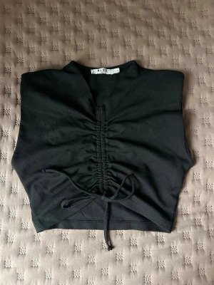 Svart croppad topp från NA-KD - Svart croppad topp från NA-KD med snörning framtill som ger en snygg rynkad effekt. Toppen är ärmlös och har en v-ringad hals. Perfekt att styla med jeans eller kjol.
