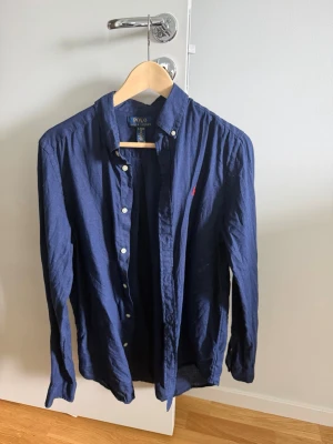 Mörkblå skjorta från Polo Ralph Lauren - Klassisk mörkblå skjorta från Polo Ralph Lauren med röd broderad logga på bröstet. Skjortan har lång ärm, knappar framtill och button-down krage. Tillverkad i mjukt bomullstyg som känns skönt mot huden. Perfekt för en stilren och avslappnad look.