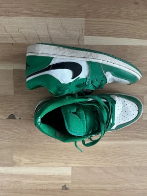 Gröna och vita Air Jordan 1 Mid - Snygga gröna och vita Air Jordan 1 Mid sneakers med svart swoosh. Skorna har klassisk hög siluett, perforerad tåbox och gröna skosnören. Tillverkade i läder med textildetaljer och ikonisk Jumpman-logga på plösen. Perfekta för dig som vill sticka ut med streetwear-stil.
