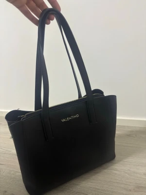 Svart handväska från Valentino - Stilren svart handväska från Valentino med två långa axelband och guldfärgad dragkedja. Väskan har en klassisk, fyrkantig form och är tillverkad i slätt material med diskret logga framtill. Perfekt för dig som gillar enkel och elegant design.