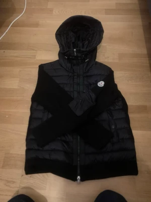 Svart dunväst med huva från Moncler - Snygg svart dunväst från Moncler med huva och tvåvägsdragkedja. Västen har ett quiltat mönster, Moncler-logga på bröstet och ribbade detaljer vid ärmöppning och nederkant. Perfekt för lager-på-lager och streetstyle.