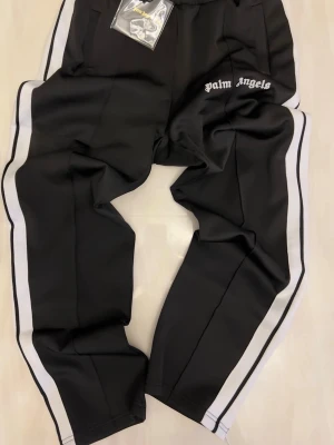 Palm Angels svarta mjukisbyxor - Svarta mjukisbyxor från Palm Angels med vita sidoränder och broderad logga på vänster höft. Byxorna har resår i midjan och raka ben. Perfekta för en avslappnad och trendig streetwear-look.De är använda men ser ändå bra ut.