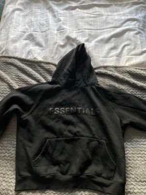 Svart Essentials hoodie med huva - Svart hoodie från Essentials med stor huva och känguruficka framtill. Snyggt broderad logga på bröstet och en liten Essentials-detalj på huvan. Perfekt för en chill och stilren look.