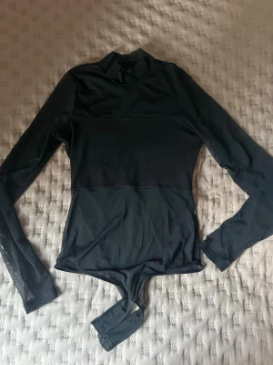 Svart långärmad body med hög krage - Snygg svart body med lång ärm och hög krage. Tillverkad i ett stretchigt material som sitter tajt mot kroppen. Perfekt att styla med jeans eller kjol för en trendig look.