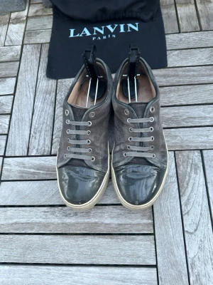 Grå sneakers från Lanvin med glansig tå - Snygga grå sneakers från Lanvin med ovandel i mocka och en glansig, lackad tå. Skorna har vita sulor och klassisk snörning. Perfekta för dig som gillar en stilren men ändå unik look. Uk 6 vilket egentligen är 40 men passar mer 41