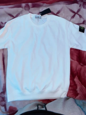 Vit sweatshirt från Stone Island - Säljer en clean vit sweatshirt från Stone Island med den klassiska svarta logotypen på ärmen. Tröjan har rund halsringning och ribbade muddar vid ärmslut och nederkant. Perfekt för dig som gillar streetwear och enkel stil.