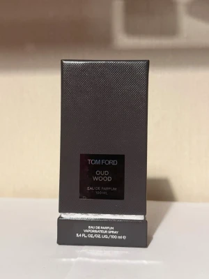 Tom Ford Oud Wood Eau de Parfum 100ml - Tom Ford Oud Wood Eau de Parfum i en elegant mörkgrå, rektangulär kartong med subtil textur. Flaskan rymmer 100 ml och doften är känd för sina lyxiga, träiga och orientaliska toner. Perfekt för dig som gillar exklusiva och unika parfymer.