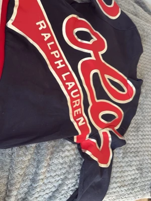 Svart och röd t-shirt från Ralph Lauren - Cool svart t-shirt från Ralph Lauren med stora röda och vita tryckdetaljer samt logga över bröstet. Tillverkad i mjuk bomull och har en klassisk passform med korta ärmar. Perfekt för dig som gillar streetstyle och vill sticka ut.