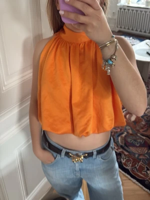 Orange halterneck topp med knyt - Snygg orange halterneck topp med hög krage och knyt i nacken. Toppen är croppad och har en lös passform med lätt glansigt tyg. Perfekt för dig som vill sticka ut med färg och stil.