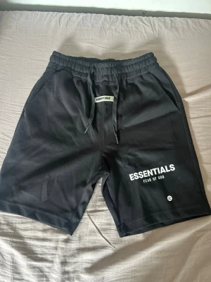 Svarta shorts från Essentials - Svarta shorts från Essentials Fear of God med vit logga på vänster ben. De har elastisk midja med snörning och är tillverkade i mjukt material som känns skönt mot huden. Perfekta för en avslappnad och trendig stil.