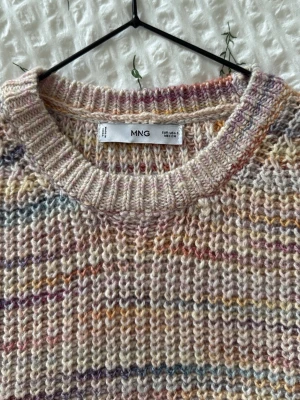 Stickad väst i pastell från MNG - Säljer en stickad väst från MNG i mjuka pastellfärger med inslag av lila, rosa, gul och blå. Västen har rund halsringning och ribbade kanter. Perfekt att styla över en skjorta eller t-shirt för en trendig lager-på-lager-look.