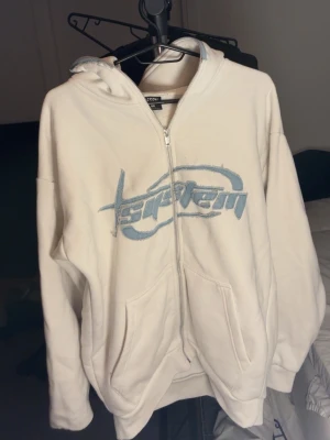 Vit tracksuit - Vit hoodie med dragkedja från System, har blått broderat tryck på bröstet. Utrustad med huva och två stora fickor framtill. Perfekt för en chill och avslappnad stil. Materialet känns mjukt och bekvämt. 