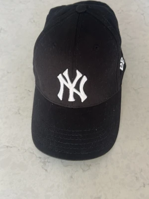 Svart New York Yankees keps - Snygg svart keps från New Era med klassiska New York Yankees-loggan broderad i vitt framtill. Kepsen har böjd skärm och MLB-logga baktill. Tillverkad i bomull och har justerbar passform. Perfekt för dig som gillar streetwear och sportig stil.