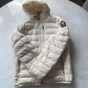 Vit dunjacka från Parajumpers YS - Säljer en superlätt, vit dunjacka från Parajumpers med huva och dragkedja. Jackan har quiltad design, bröstficka med dragkedja och klassisk PJS-logga på ärmen. Perfekt för kalla dagar när du vill hålla stilen och värmen.