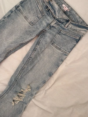 Ljusblå bootcut jeans med slitningar - Säljer ett par ljusblå bootcut jeans med slitningar på båda knäna och stora framfickor. Jeansen har en tvättad look och vida ben. Perfekta för dig som gillar en avslappnad och trendig stil.