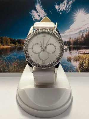 Vit klocka med strassdetaljer - Stilren klocka med vit läderrem och silverfärgat ur med glittrande strass runt boetten. Urtavlan är vit med dekorativa stenar och romerska siffror. Perfekt accessoar för att lyfta din outfit. Nytt batteri, säljes i befintligt skick enligt bilderna!