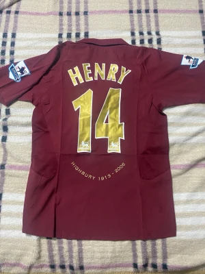 Arsenal Highbury tröja Henry 14 - Säljer en vinröd Arsenal Highbury 1913-2006 matchtröja med Henry 14 i guld på ryggen. Tröjan har korta ärmar, Premier League-patchar på ärmarna, klubbmärke och Nike-logga framtill samt O2 som sponsor. Perfekt för fotbollsfans och samlare. Priset är ej hugget i sten! Helt ny med tagg!