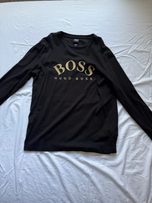 Svart långärmad tröja Hugo Boss - Svart långärmad topp från Hugo Boss med guldig BOSS-logga framtill. Klassisk rund halsringning och stilren design, perfekt för dig som gillar snygga och enkla plagg med exklusiv känsla. Tillverkad i mjuk bomull för skön komfort. 