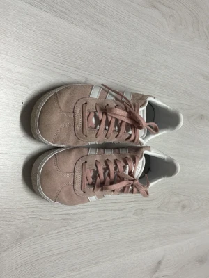Adidas Gazelle rosa sneakers - Snygga Adidas Gazelle sneakers i rosa mocka med vita detaljer och klassiska tre ränder på sidorna. Skon har lågt skaft, rosa snören och guldtexten 'GAZELLE' på sidan. Perfekt för en som gillar stilrena och trendiga sneakers.