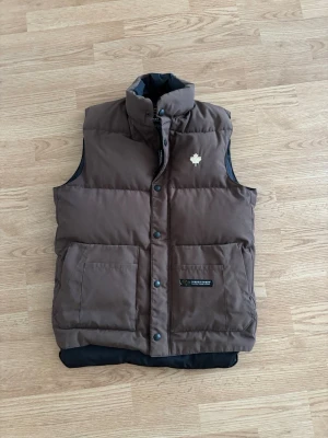 Brun dunväst från Canada Goose - Snygg brun dunväst från Canada Goose med hög krage och två stora fickor framtill. Västen har en vit lönnlöv-detalj på bröstet och svart innerfoder. Perfekt för lager-på-lager och streetstyle.