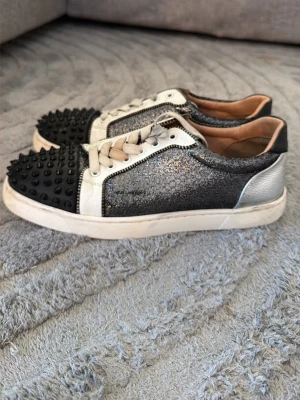 Christian Louboutin Sneakers - Säljer ett par unika sneakers från Christian Louboutin med svart glitter, silvriga detaljer och svarta nitar på tån. Skorna har vita snören, vit sula och svart hälparti. Materialmix av läder och textil ger en lyxig känsla.