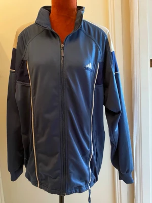Blå Adidas träningsjacka L - Snygg blå träningsjacka från Adidas med dragkedja framtill, vita detaljer och logga på bröstet. Jackan har hög krage, muddar vid ärmslut och dragsko nertill. Perfekt för sport eller chill. Materialet är troligtvis polyester.