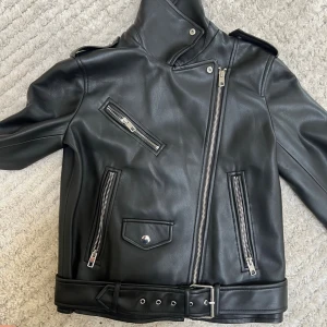 Svart klassisk skinnjacka med bälte - Snygg svart skinnjacka i bikerstil med sned dragkedja, tre framfickor med dragkedjor och dekorativt bälte i midjan. Jackan har bred krage med tryckknapp och coola detaljer i silver. Perfekt för dig som vill ha en edgy look.  OBS: Lite slitage vid kragen (se bild)