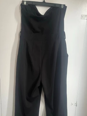 Svart axelbandslös jumpsuit - Snygg svart axelbandslös jumpsuit med hjärtformad urringning och vida ben. Perfekt för dig som vill ha en stilren och trendig look. Tillverkad i ett mjukt och följsamt tyg som ger en skön passform. Lite skadad nere vid byxbenet som visas på bild. 