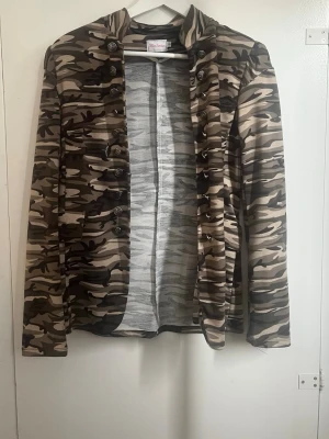 Kavaj med militärmönster och knappar - Snygg kavaj i camouflage-mönster med dekorativa knappar framtill. Rak passform och lång ärm. Perfekt för en cool och avslappnad look. Färgskalan går i grönt, beige och brunt.
