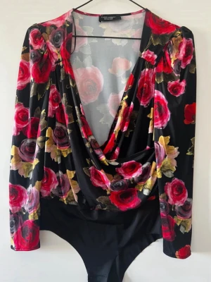 Blommig body med djup urringning - Säljer en snygg body från Zara med svart botten och stora röda, rosa och lila rosor. Bodyn har lång ärm och en djup draperad urringning framtill. Materialet är stretchigt och mjukt, perfekt för att skapa en cool look. Oanvänd 