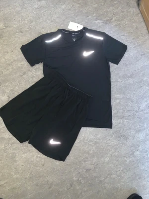 Svart träningsset från Nike - Snyggt svart träningsset från Nike med t-shirt och shorts. Båda plaggen har reflekterande Nike-loggor och är gjorda i ett lätt och snabbtorkande material som passar perfekt för träning. T-shirten har rund hals och korta ärmar, shortsen har resår i midjan.
