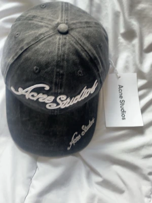 Svart keps från Acne Studios - Snygg svart keps från Acne Studios med vit broderad logga framtill och på skärmen. Kepsen har klassisk sexpaneldesign och justerbar rem baktill. Tillverkad i bomull med en tvättad vintage-look. Perfekt accessoar för en trendig streetstyle.