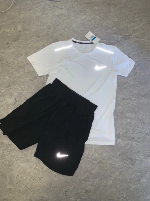 Nike träningsset t-shirt och shorts - Snyggt träningsset från Nike med en vit t-shirt och svarta shorts. Båda plaggen har den klassiska Nike-loggan i vitt. T-shirten är kortärmad och har rund hals, shortsen har resår i midjan och dragsko. Perfekt för gymmet eller löpning.