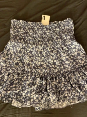 Blommig kortkjol med volang - Supersöt kortkjol med blommigt mönster i blått och vitt. Kjolen har en smockad midja och volang nertill som ger en lekfull look. Perfekt för dig som gillar romantisk och trendig stil.