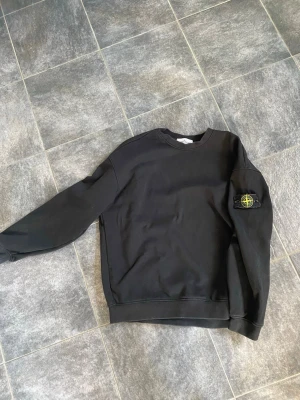Svart sweatshirt från Stone Island - Svart sweatshirt från Stone Island med klassisk rund hals och ikonisk logotyp-patch på vänster ärm. Tröjan har en avslappnad passform och är tillverkad i mjukt bomullsmaterial. Perfekt för en stilren och trendig look.