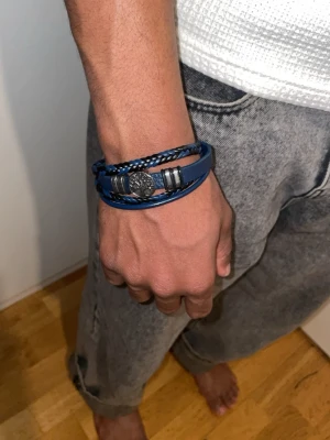 Blått läderarmband med trädberlock - Snyggt armband i blått och läder med flätade detaljer och en silverfärgad berlock med trädmotiv. Magnetlås i metall och flera lager ger en cool och modern look. Perfekt accessoar för att lyfta din outfit.