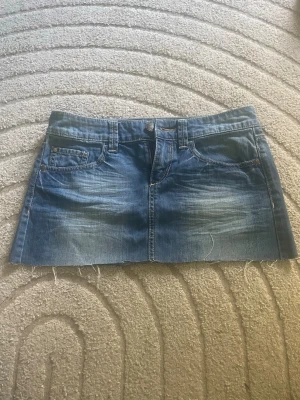 Blå jeanskjol med rå kant - Snygg blå jeanskjol med klassisk femficksdesign och rå, avklippt kant nertill. Kjolen har slitna detaljer framtill och dekorativa sömmar på bakfickorna. Perfekt för en avslappnad och trendig look. midjemått 38cm längd 28cm