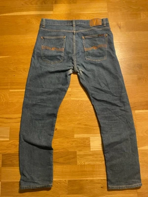 Blå jeans från Nudie Jeans - Säljer ett par klassiska blå jeans från Nudie Jeans med raka ben och femficksmodell. Jeansen har tydliga sömmar på bakfickorna och en patch i läderimitation vid linningen. Perfekta för en avslappnad och stilren look. Slitage som lagats i skrevet men syns inte när man har dom på sig.