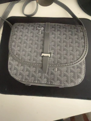 Grå axelväska från Hm - Snygg grå axelväska från Hm med det klassiska mönstret i vitt och grått. Väskan har justerbar axelrem och ett lock med spänne framtill. Tillverkad i canvas med läderdetaljer. Perfekt för dig som gillar stilrena accessoarer.