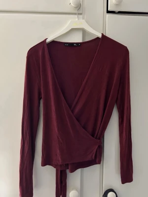 Vinröd topp med knyt - Vinröd omlottopp med långa ärmar och knyt i midjan. Djup v-ringning framtill och mjukt, lätt material som ger en snygg och avslappnad look. Perfekt att styla med jeans eller kjol.