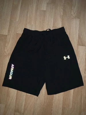 Svarta shorts från Under Armour - Svarta shorts från Under Armour med snörning i midjan och logga på ena benet. Shortsen har även texten 'ARMOUR' i regnbågsfärger längs benet. Perfekta för träning eller chill.