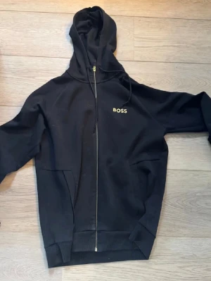 Svart zip-hoodie från BOSS - Svart hoodie med dragkedja från BOSS. Klassisk design med huva och snörning, två fickor framtill och diskret BOSS-logga på bröstet. Perfekt för en chill och stilren look.