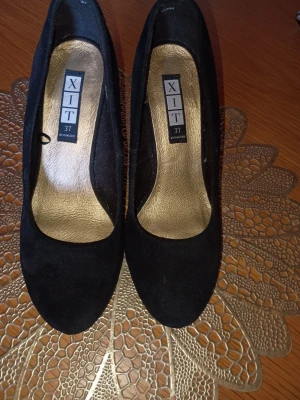 Svarta pumps från XIT i mockaimitation - Stilrena svarta pumps från XIT i storlek 37. Skorna har rundad tå och är tillverkade i mockaimitation med guldfärgad innersula. Perfekta för dig som vill ha en klassisk och enkel look med en touch av elegans.